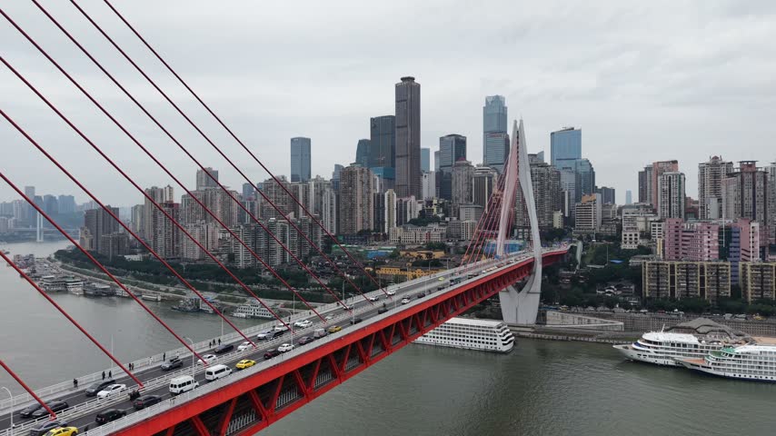 Descending aerial fly past along a red cable stayed bridge from above down to the river, revealing a large cruise ship moored below the deck, overcast haze, Chongqing, China.