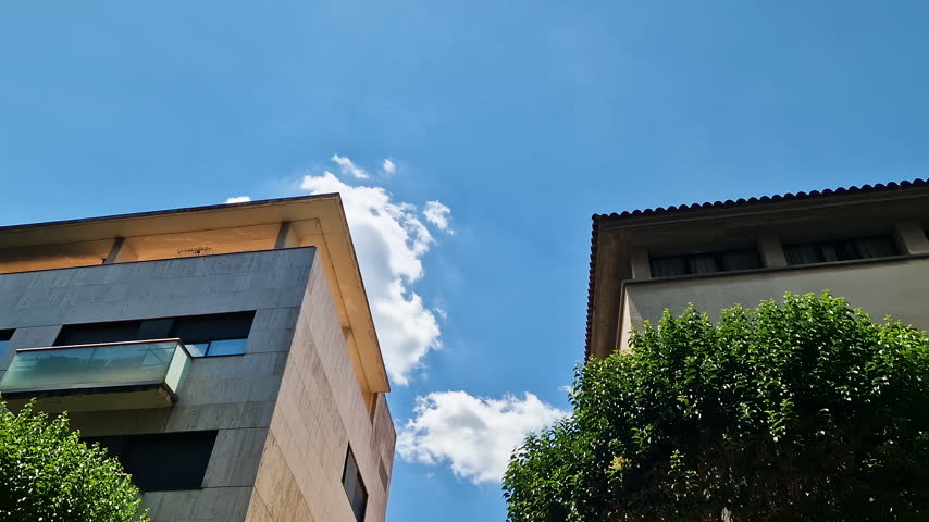 POV looking up at apartment buildings and green trees against a bright blue summer sky in Spain
