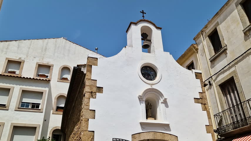POV looking up at a historic white church facade with a bell tower under a clear blue sky in Spain