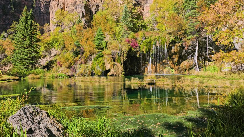 Tranquil lakeside landscape showing small waterfalls descending from moss covered rocks, surrounded by autumn leaves, rugged cliff textures, and calm water with gentle ripples.