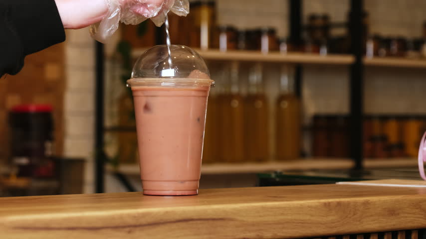 Barista hand with bar spoon stirs iced coffee in plastic cup at counter closeup. Barkeeper makes cold cocktail with ice cubes in coffeeshop