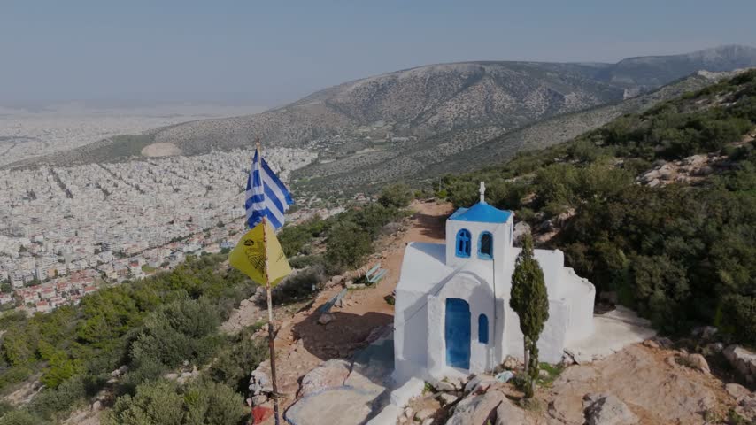 Cinematic aerial drone view of a traditional white and blue Greek chapel on a rocky mountainside overlooking Athens, Greece. The drone flies past the chapel revealing the dense cityscape, surrounding mountains and Mediterranean landscape.