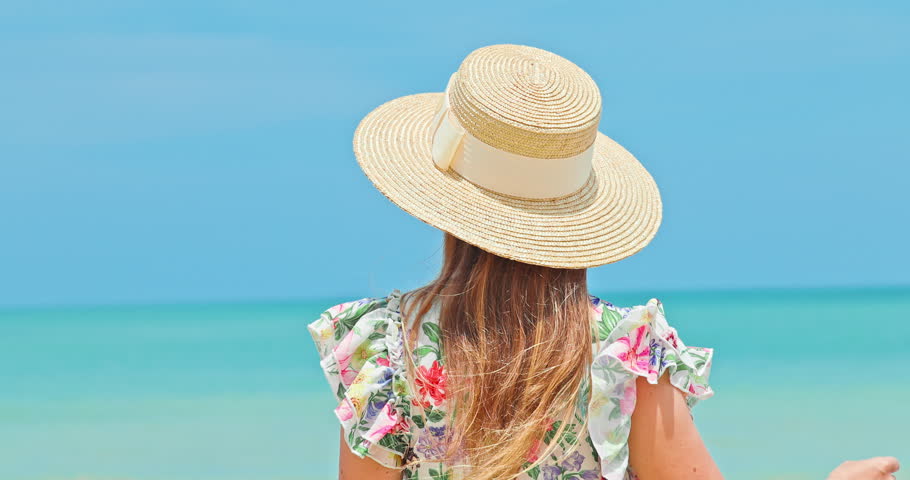 Young woman wearing straw hat and floral dress looking at turquoise ocean. Rear view of female traveler enjoying sunny tropical beach. Summer holiday travel concept.