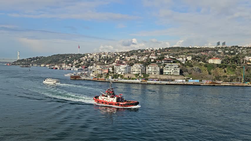 Red boat navigates busy waterway. Cityscape lines distant shoreline. Sky shows scattered clouds above. Boats move through calm blue waters. Water ripples trail behind vessels in Istanbul, Turkey