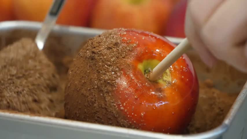 close-up shot captures a hand holding a stick inserted into a bright, glossy red apple coated in a smooth, translucent caramel or candy layer.