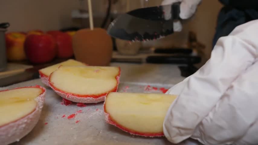 A close-up shot of a person wearing white protective gloves using a sharp knife to carefully slice a fresh apple coated in a bright red candy shell on a light gray cutting board.