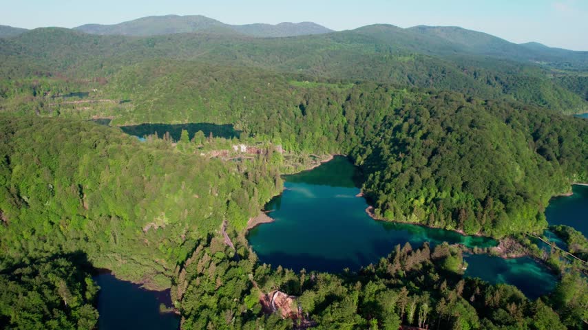 Dynamic aerial view of plitvice lakes national park, showcasing numerous turquoise lakes, stunning waterfalls, and dense green forests under bright summer sky, beautiful untouched nature scenery.