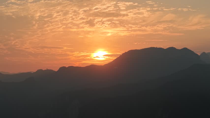 Dramatic sunrise over the Taihang Mountains. The golden sun rises behind sharp mountain silhouettes, illuminating a textured orange sky. Aerial view of Heimaogou Grand Canyon, China.