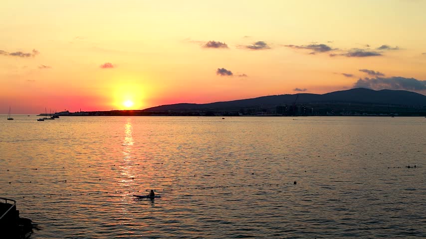 Sunset over calm water with boats sailing, silhouetted trees framing the scene, and distant hills creating a serene landscape in a coastal region