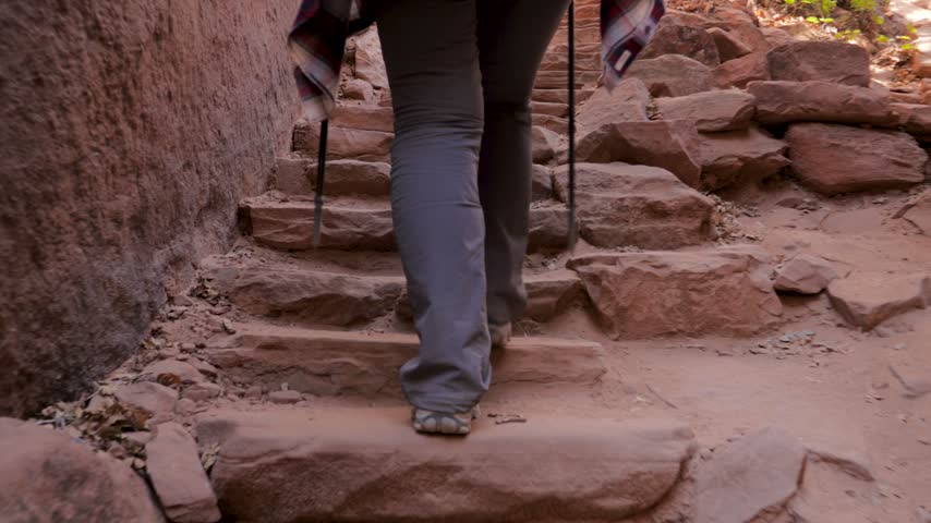 Close up view of trekker feet and legs rises upward steep, rugged stone staircase carved into red rock canyon, using trekking poles for stability during an adventurous hike