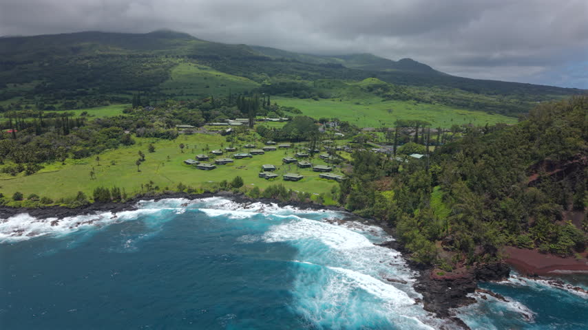 Aerial view of Kaihalulu Beach on Maui Hawaii island with turquoise surf breaking on black lava rocks green pastures palm trees and low cloud covered volcanic slopes rising above the tropical coast.
