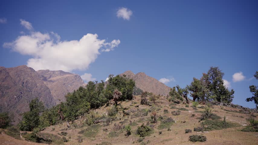 Puffy clouds drift over a jagged mountain ridge above a forested hilltop lined with an old dry stone wall.