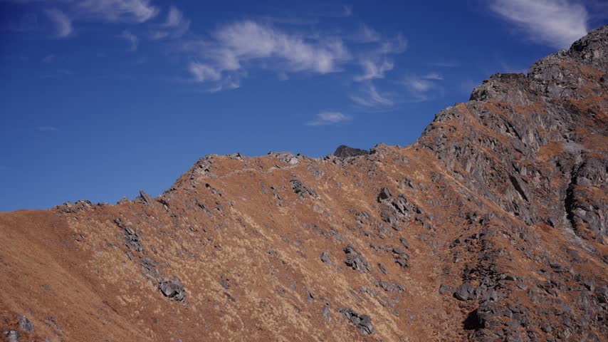 A golden ridge trail winds toward a rocky summit beneath streaking wispy clouds, capturing the dramatic scale of far west Nepal