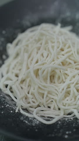 Vertical close-up of pale thin noodles piled on a black plate with white flour scattered around them.