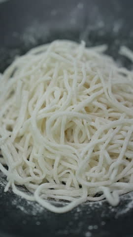 Vertical close-up of pale thin noodles piled on a black plate with white flour scattered around them.