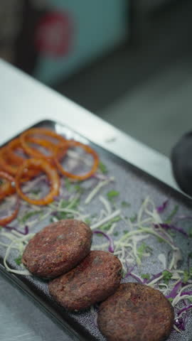 Vertical close-up of a chef arranging browned round patties over shredded vegetables on a rectangular platter beside tomato slices.