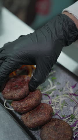 Vertical close-up of a chef arranging browned round patties over shredded vegetables on a rectangular platter beside tomato slices.