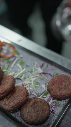 Vertical close-up of a chef arranging browned round patties over shredded vegetables on a rectangular platter beside tomato slices.