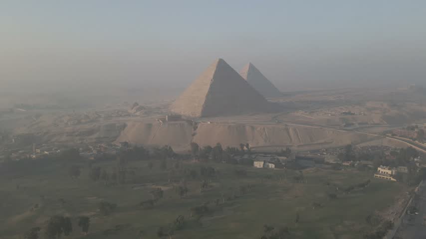 Aerial drone footage approaching two pyramids of Giza, with lush green trees and cultivated land visible in the foreground contrasting the desert landscape, Egypt.