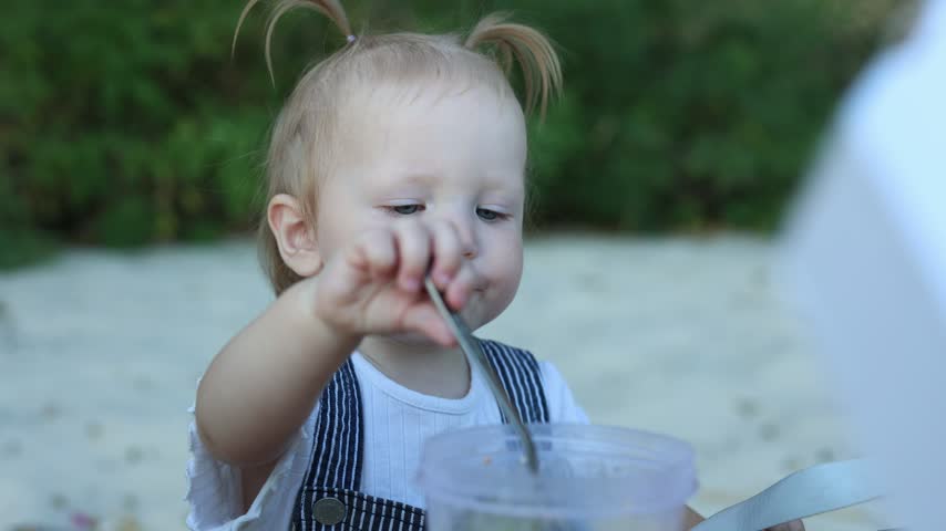 A young child with blonde pigtails and blue eyes enjoys a meal with a spoon, showing early signs of independence and curiosity