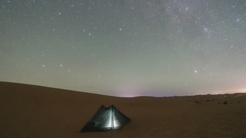 A lit camping tent sits on a sand dune beneath a clear night sky filled with stars and the Milky Way.