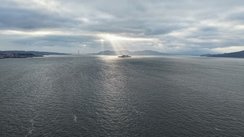 Aerial tracking view moving forward over open bay water toward Alcatraz Island, dramatically lit by breaking sun rays under a heavy overcast storm sky