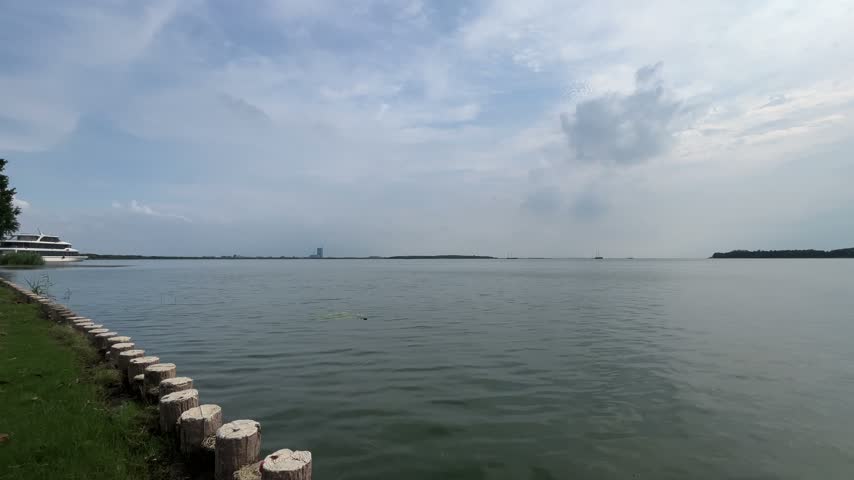 Static wide shot of calm lake waters with gentle ripples on a cloudy daylight afternoon at Suzhou Bay. Wooden piling posts border the grassy shoreline in the foreground, with a large boat docked on the left under a soft overcast sky. Perfect background footage for nature, environmental, and travel projects.