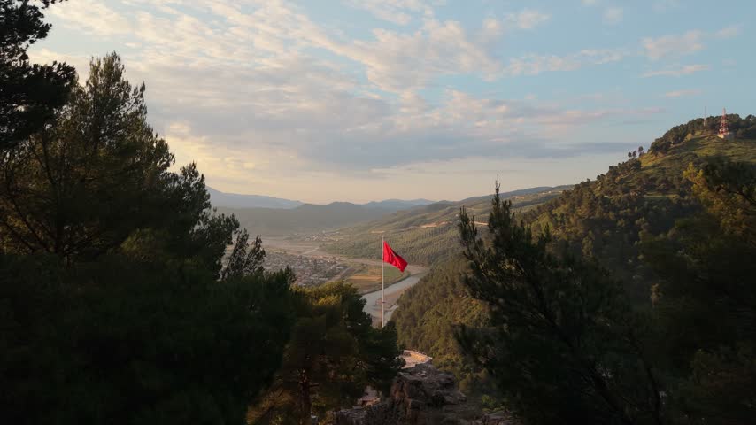Aerial view of a red flag waving over the Osum river valley surrounded by mountains and pine trees in Berat, Berat County, Albania.