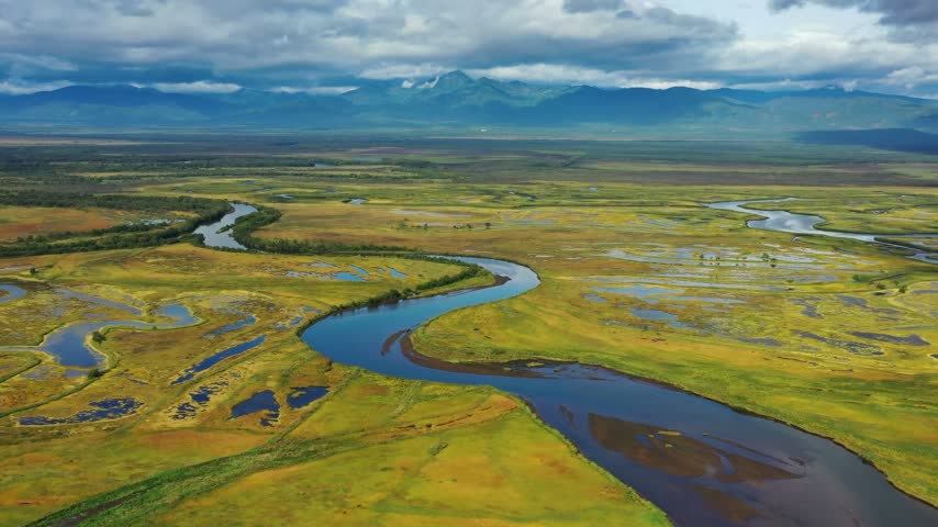 Aerial view of a winding river flowing through the marshy landscape with distant mountains under a cloudy sky, Kamchatka, Russia.