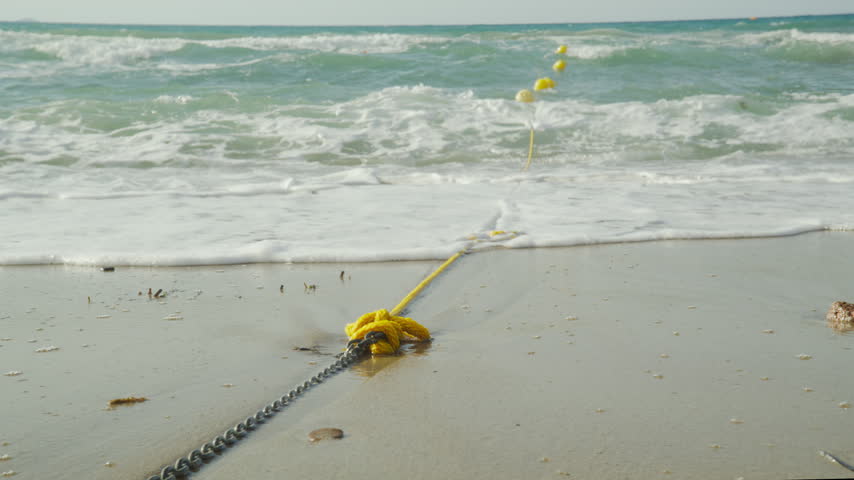 A yellow rope and metal chain lie on a sandy beach as rough ocean waves wash ashore towards floating yellow buoys