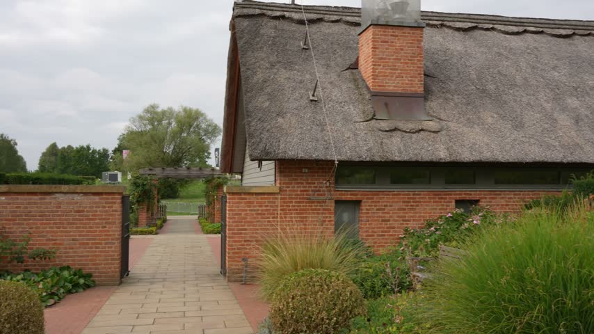 Camera pans along a brick house with a thatched roof and chimney, past a garden path and ornamental grasses, ending on tall grass.