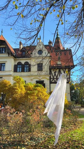 White ghost decoration hangs on tree branch near historic castle building in autumn park. Seasonal Halloween outdoor decor and spooky gothic atmosphere concept.