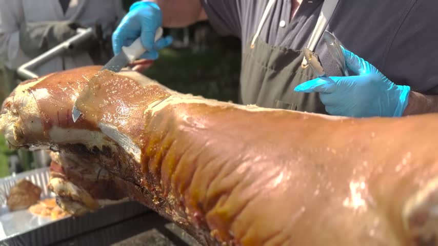 Close-up video footage of a chef carving a deliciously roasted pig with crispy skin during an outdoor barbecue event, highlighting culinary preparation.
