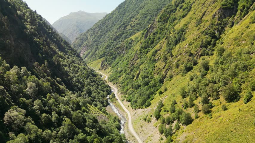 Aerial drone footage of a winding unpaved mountain road leading into a narrow gorge, surrounded by steep green slopes and dense forest.