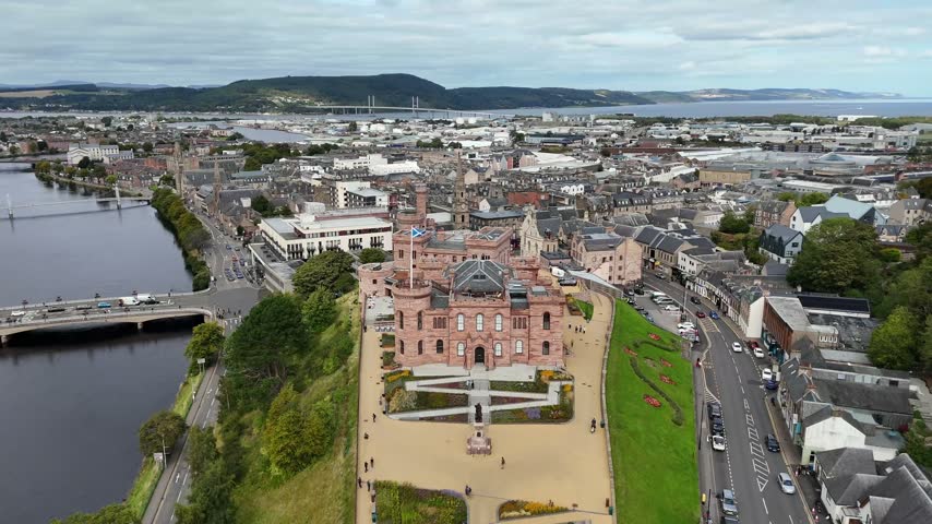 Elevated straight-on aerial drone video of Inverness Castle front facade, Scottish flag, River Ness bridge, city center buildings, and Kessock Bridge in Highland, Scotland