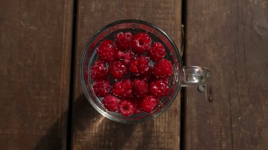 Bright red raspberries in a glass cup, viewed from above on brown wooden planks. Minimal food photography background with fresh summer berry atmosphere
