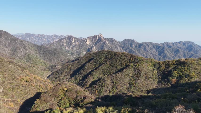 Static hovering aerial over layered mountain ridges near the Great Wall in Huairou District, autumn colored slopes with patches of green foliage extending to distant hazy peaks, Beijing, China.