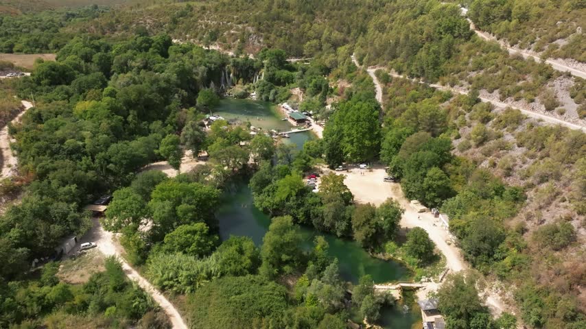 Recreation areas, parking lots and forested gorge surround the Kravica waterfall pools as a drone flies high overhead.
