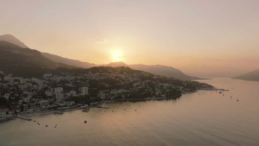 Sun dips behind layered hills as a dense coastal apartment district meets calm reflective water in this gentle drone flyover.