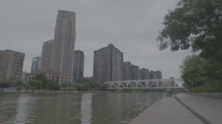 Wide static view of a city riverfront featuring modern high-rise residential buildings, a commercial skyscraper, and a white steel truss bridge under an overcast sky.