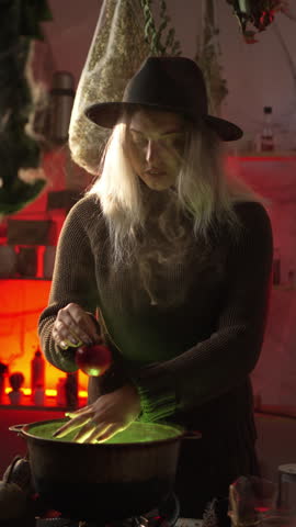 A witch brews a potion in a cauldron among dried herbs and cobwebs in an authentic home environment. She holds a red apple over a cauldron with a potion and moves it.