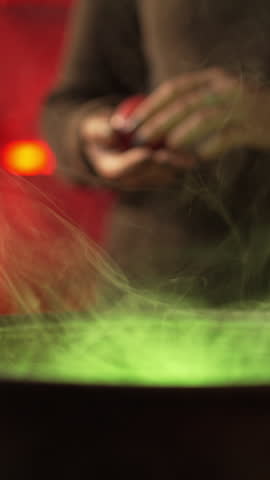 A witch brews a potion in a cauldron among dried herbs and cobwebs in an authentic home environment. She holds a red apple over a cauldron with a potion and moves it.