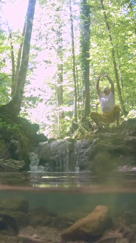 A man practices yoga on a rocky outcrop by a tranquil stream in a serene forest, surrounded by tall trees and dappled sunlight, promoting a zen-like connection with nature and a healthy lifestyle.