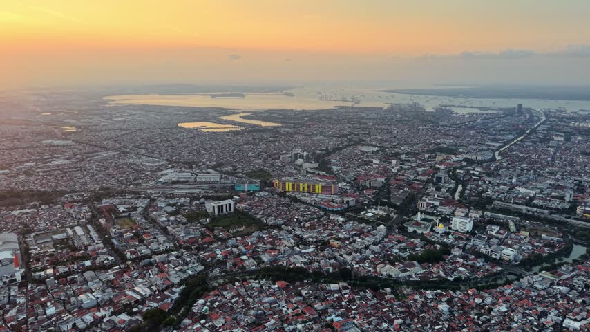 Expansive Aerial view of Surabaya at sunset showing dense neighborhoods, waterways, ports, and glowing coastal horizon