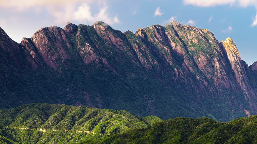 Massive granite mountain ridge covered in green vegetation stands against a blue sky with soft clouds in bright daylight.