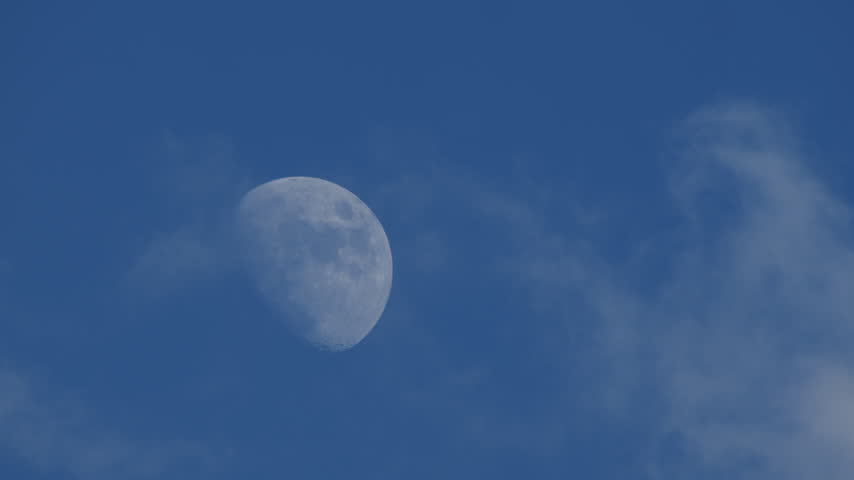 A half Moon visible in the blue sky during late afternoon or evening daylight, with soft clouds floating past it. 