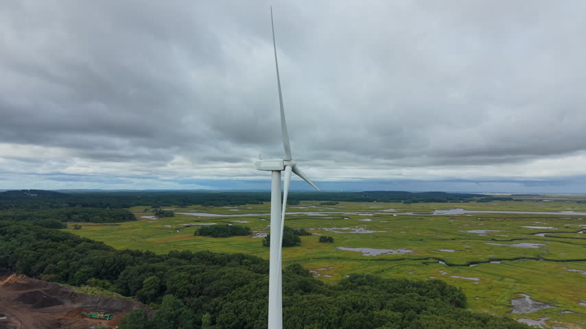 White wind turbine towering over Great Marsh wetlands in Ipswich Massachusetts, winding tidal creeks, salt marsh, dense green trees, coastal landscape under cloudy sky. Aerial drone view.