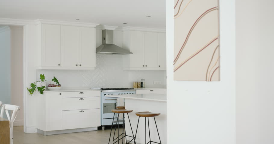 Camera is beginning to pan right and zoom out, showing bright kitchen with island, stools, sink. Sunlit, minimalist, modern, white, cabinetry, countertops, shelves
