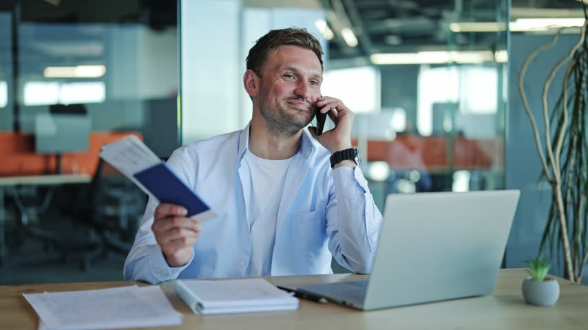 Office worker in a casual shirt discusses travel plans on his smartphone, holding a passport and plane tickets, sitting at his modern office desk with a laptop.