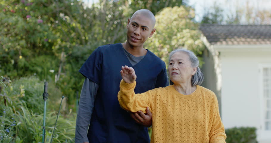 Senior Asian woman holding cane on path pointing to house, African-American navy-clad nurse guiding. Caregiver, patient, outdoor, garden, walking, dialogue, wellbeing